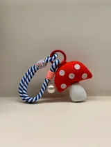 Llavero o charm para cartera con forma de hongo de tela roja con lunares blancos bordados y tallo blanco. Incluye una correa de cuerda trenzada en azul y blanco, un dije de perla y un mosquetón metálico rojo.