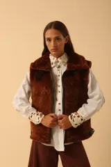 Chaleco de piel sintética en color chocolate, con cierre frontal y detalles de cuero en los bordes. Presenta un diseño de cuello solapa y ajuste lateral con hebillas.