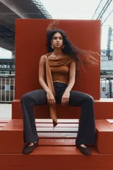 Top color marrón de punto elástico, con un hombro descubierto y una tira larga que cae desde el hombro. Presenta fruncidos en los laterales para un efecto drapeado.