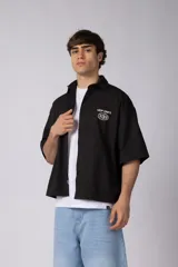 Camisa de manga corta negra, de corte boxy, con cuello camisero, cierre frontal de botones y bordado circular en el pecho.