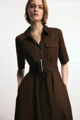 Vestido midi de cuello solapa y escote en pico, con manga corta con vuelta. Presenta bolsillos laterales ocultos, solapas decorativas en el pecho y cierre frontal con botones. Incluye cinturón de lazada en el mismo tejido.
