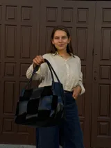 Cartera de hombro confeccionada en cuero y gamuza con diseño de damero, combinando paneles de texturas lisas y aterciopeladas. Presenta una manija trenzada y detalles de costuras artesanales en los laterales.