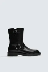 Bota de media caña en color negro, con diseño de horma cuadrada y suela con ligero tacón. Presenta dos cintas ajustables con hebillas metálicas, una en la parte superior de la caña y otra en la zona del empeine.