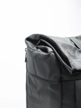 Mochila negra con cierre superior enrollable y hebilla de plástico. Cuenta con un bolsillo frontal con el texto "FORUM INDUSTRIES CO." y un parche en forma de diamante en la parte inferior. Tiene un bolsillo lateral con cierre.
