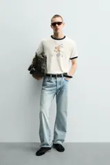Camiseta de algodón color crudo con cuello y bordes de mangas en contraste negro. Presenta un estampado frontal de estilo vintage con el personaje Striker, mascota de la Copa Mundial de la FIFA 1994, jugando al fútbol.