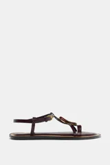 Sandalia plana de cuero color marrón oscuro (bordó/caoba) con tira en T y hebilla ajustable en el tobillo. Presenta un aplique decorativo curvo en tono ámbar/marrón rojizo con borde dorado en la tira del empeine.