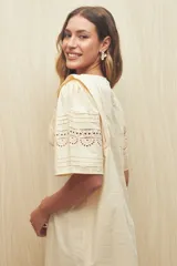 Vestido corto color crema, de corte recto y cuello redondo. Presenta mangas cortas amplias con calados geométricos en el ruedo.