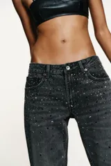 Jeans de tiro bajo y corte recto, confeccionados en denim color gris oscuro con efecto lavado. Presentan cinco bolsillos y están decorados con aplicaciones de strass multicolor en diferentes formas y tamaños en la parte frontal. Cierre frontal con cremallera y botón metálico.