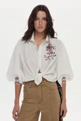 Blusa de mezcla de algodón y lino color blanco, con mangas abullonadas y cierre frontal de botones. Presenta detalles de bordado geométrico a cuadros en el cuello y los puños, y un gran bordado floral en el pecho.