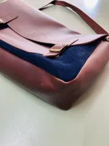 Maletín tipo cartero con solapa de cuero marrón y cuerpo de gamuza azul. Cuenta con correas ajustables con hebillas metálicas, asa superior y correa larga para llevar al hombro.