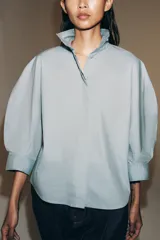 Camisa de algodón con cuello alto y mangas abullonadas por debajo del codo. Presenta cierre frontal con botones ocultos bajo solapa.