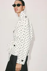 Cazadora estilo trench corta confeccionada en lino, con diseño de lunares negros sobre fondo blanco. Cuenta con cuello solapa, manga larga con trabillas en los puños, cierre frontal cruzado con botones negros y cinturón ajustable del mismo tejido.
