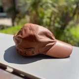 Gorra de cuero metalizado dorado, con ojales metálicos decorativos y etiqueta de marca aplicada en el lateral.