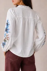 Camisa blanca de manga larga con bordados florales en tono rojo y verde en el frente y las mangas. Presenta cuello mao con escote en V y botones frontales.