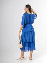 Vestido midi de seda italiana color azul, con escote en V, mangas murciélago amplias, falda de tres volados y cintura elastizada con lazo ajustable al tono y hebilla circular dorada.