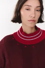 Buzo de tejido de punto con diseño de trenzas, color crema con detalles en marrón en cuello, puños y ruedo. Presenta líneas finas en contraste y pequeñas costuras decorativas en forma de cruz.
