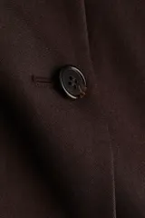 Campera corta de sarga color marrón oscuro, con cuello alto, cierre frontal con botones y bolsillos laterales. Presenta mangas largas abullonadas con fruncidos en los hombros y puños ajustados.
