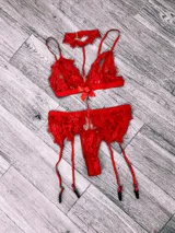 Conjunto de lencería rojo de encaje floral, compuesto por corpiño triangular con breteles finos y moño en el centro, tanga less y portaligas con tiras ajustables. Incluye gargantilla de encaje con cierre trasero.