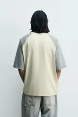 Camiseta de corte relajado con mangas ranglán en color gris y cuerpo en color beige. Presenta cuello redondo, costuras a contraste y detalles de parches y bordados en el pecho.
