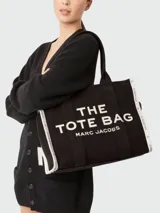 Bolso tote de lona color beige con estampado frontal en negro que dice 'THE TOTE BAG MARC JACOBS'. Presenta bordes laterales en color negro con detalles de texto y doble asa superior.
