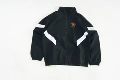 Campera deportiva negra de nylon liviano con paneles diagonales blancos en contraste. Presenta un estampado frontal discreto y un gran gráfico en la espalda que incluye una silla naranja con un gato blanco durmiendo y la frase "Sin siesta no hay fiesta".