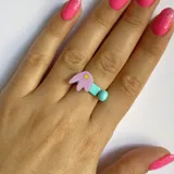 Anillo ajustable de metal con aplique de figura abstracta en color lila y celeste, con detalle de punto amarillo.