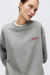 Buzo oversized de felpa color gris melange, con cuello redondo y hombros caídos. Cuenta con un pequeño bordado rojo con texto en el pecho y puños y bajo con ribete acanalado.