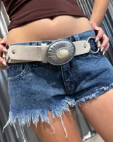 Cinturón de cuero sintético en color beige, con diseño de hebilla metálica grande de estilo western, decorada con un detalle central de piedra clara y acabado plateado envejecido. Presenta argollas metálicas laterales y remaches decorativos.