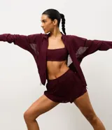 Campera deportiva tipo rompevientos color bordó, con cierre frontal, cuello alto y mangas largas. Presenta paneles laterales de tul para mayor ventilación y cordón ajustable en la cintura.