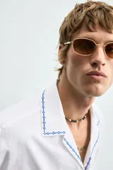 Camisa blanca de algodón con cuello bowling y manga corta, corte relajado. Presenta bordados a contraste en azul, rojo y amarillo que simulan elementos de cocina y artesanía, además de ribetes de pespunte azul en cuello, puños y abertura frontal.