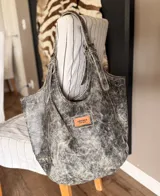 Cartera tote bag tipo shopper artesanal, elaborada en cuero con efecto desgastado color gris y negro. Cuenta con amplias asas de hombro regulables, tachas decorativas en la base y un parche frontal de cuero con el logo de la marca.