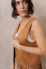 Chaleco de efecto gamuza en tono camel, sin mangas, con escote en V y diseño abierto. Presenta detalles de tachas metálicas, entramado artesanal en el frente y flecos largos en el ruedo.