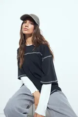 Remera de manga corta color marrón con efecto desgastado, corte oversize y costuras a la vista en color blanco. Presenta cuello redondo con cuello polo blanco superpuesto y mangas largas a rayas horizontales marrones y blancas por debajo.