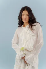 Camisa de estilo romántico confeccionada en algodón, con cuello alto, volados frontales y mangas largas con puños acampanados también decorados con volados.