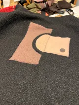 Campera de textura tipo bouclé en color negro, con diseño de collage frontal compuesto por apliques de diferentes texturas y tonos tierra, incluyendo un bolsillo cuadrado con botón metálico.