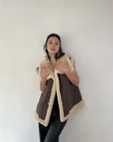 Chaleco estilo gamulán de cuero sintético marrón con interior y bordes de corderito sintético color crema. Presenta cierre frontal con botones a presión y bolsillos laterales.