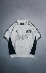 Chomba de punto estilo deportivo, color blanco con detalles en negro. Presenta cuello polo con rayas, mangas cortas con franjas laterales, y diseño frontal con el número 90 y la palabra Ravers bordada.