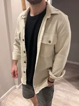 Sobre camisa de jean color arena, de corte recto, con cuello clásico, cierre frontal con botones, mangas largas con puños abotonados, bolsillos con solapa abotonada en el pecho y ruedo recto.