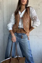 Chaleco de gamuza color camel con bordados decorativos en color blanco y cierre frontal con cordones y apliques metálicos.