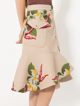 Pollera midi de gabardina color beige con estampado de flores y bordados. Presenta un cruce diagonal con botón dorado personalizado y botón interno para sostener la solapa inferior. Incluye bolsillo plaqué en delantero orientado a cintura y volado en forma de caracol en el ruedo.