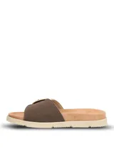Sandalias tipo slide con tira ancha en tejido texturizado color beige y detalle de hebilla circular decorativa en tono marrón. Presentan una base ergonómica con suela de poliuretano ligera y flexible.