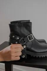 Bota corta de cuero negro con punta cuadrada, suela track y detalle de estribo desmontable con hebilla metálica.