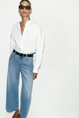 Jeans de tiro alto con pernera ancha y corte cropped. Presenta diseño de cinco bolsillos, cierre frontal con cremallera y botón, e incluye un cinturón oscuro con hebilla metálica.