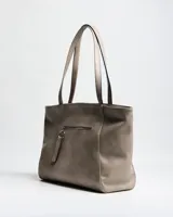 Cartera tipo tote de símil cuero color taupe, con doble asa de hombro, detalles de tachas plateadas en los laterales frontales y placa metálica con logo grabado de la marca XL Extra Large en el centro.