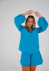 Conjunto de dos piezas compuesto por una camisa de corte relajado con mangas abullonadas, puños elásticos y cierre frontal de botones, y un short de tiro medio con cintura elástica. Ambas prendas están confeccionadas en lino liviano de color turquesa.