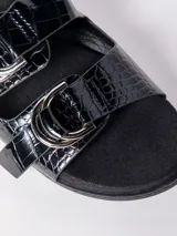 Sandalia baja negra con textura de cocodrilo (croco) en las tiras anchas. Presenta dos hebillas metálicas plateadas ajustables. La base es negra y plana.
