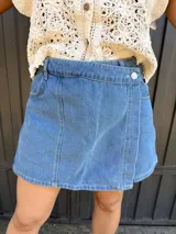 Short pollera de jean de lavado medio, con diseño cruzado asimétrico y cierre frontal con botón metálico, costuras verticales y bolsillos delanteros.