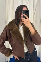 Campera bomber corta de cuero ovino marrón con detalles en napalan coñac y forro interior. Presenta cuello solapa, cierre frontal con cremallera de doble sentido, bolsillos laterales ribeteados, mangas largas con detalles de pasamanería cruzada y puños de piel sintética.