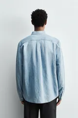 Camisa de corte regular fit confeccionada en denim ligero de algodón con efecto lavado. Presenta cuello solapa con botones, manga larga con puños abotonados, cierre frontal de botones y un bolsillo plastrón en el pecho.
