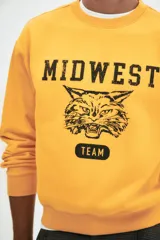 Buzo de tejido pesado de algodón color amarillo mostaza, con cuello redondo ribeteado, hombros caídos y puños y bajo en punto acanalado. Estampado frontal con el texto "MIDWEST" en mayúsculas, un dibujo de un felino rugiendo y la palabra "TEAM" en un óvalo.